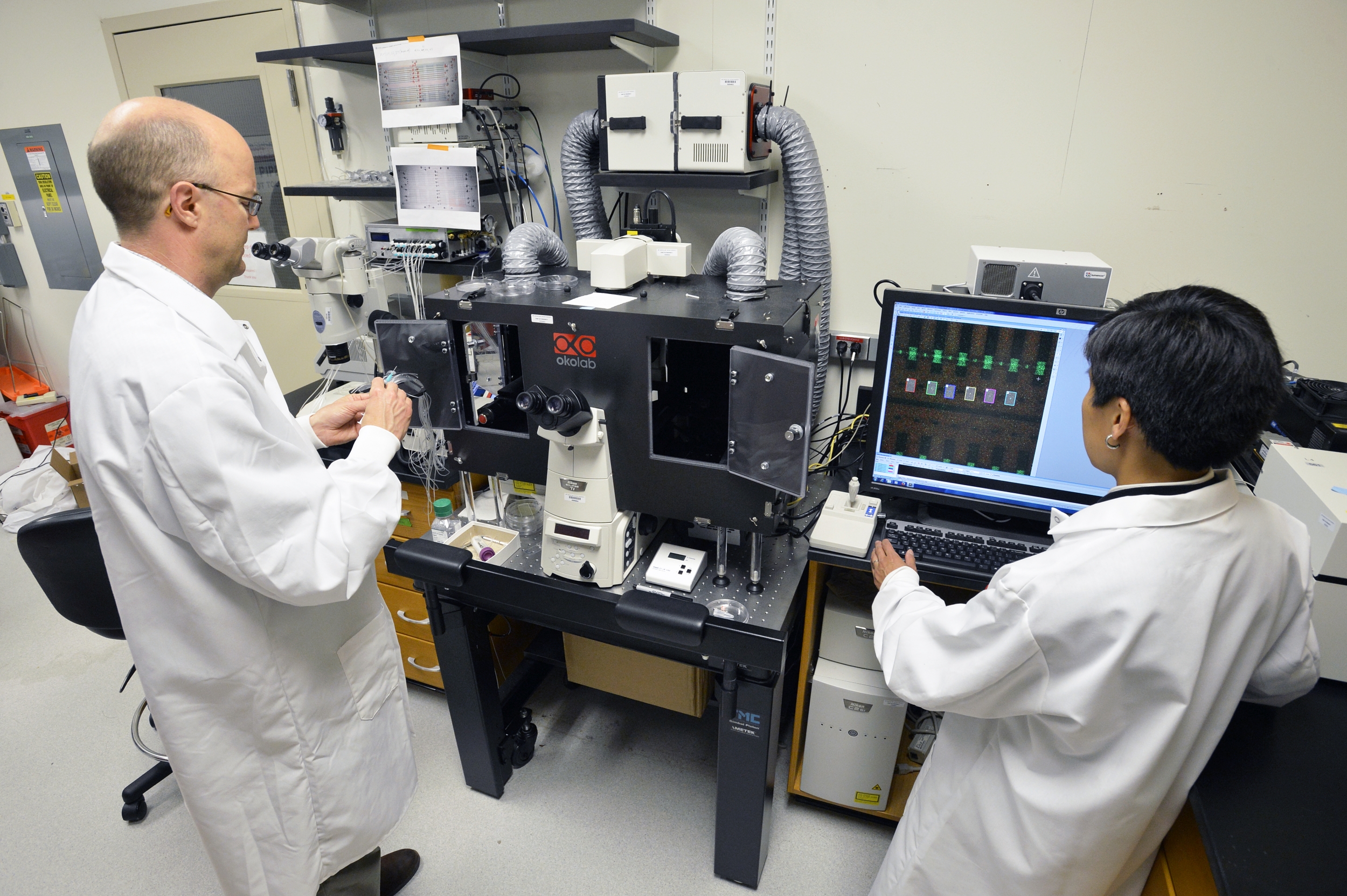 Two researchers in white lab coats stand in front of a large microscope and monitor. 