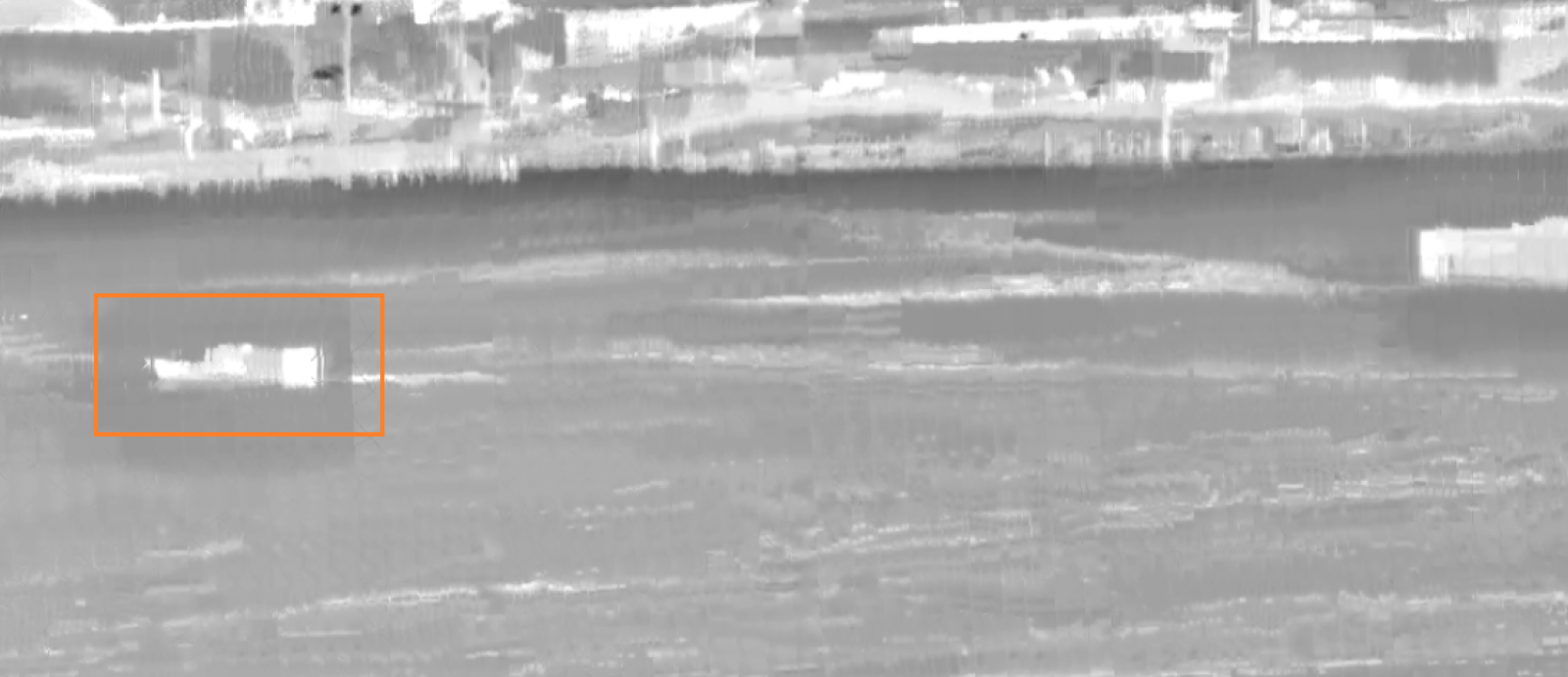 A grey-scale surveillance image of a shoreline, with a boat in the water outlined by an orange box.