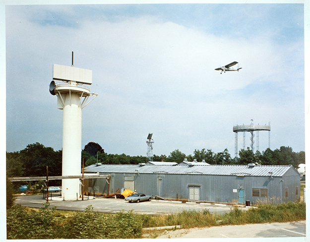 A photo of the exterior of a building, a small radar tower, and an aircraft in the background.