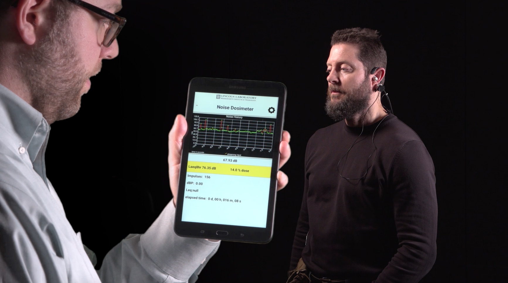 A researcher holds a tablet showing the MNOISE app while a user wears the MNOSIE earpiece.