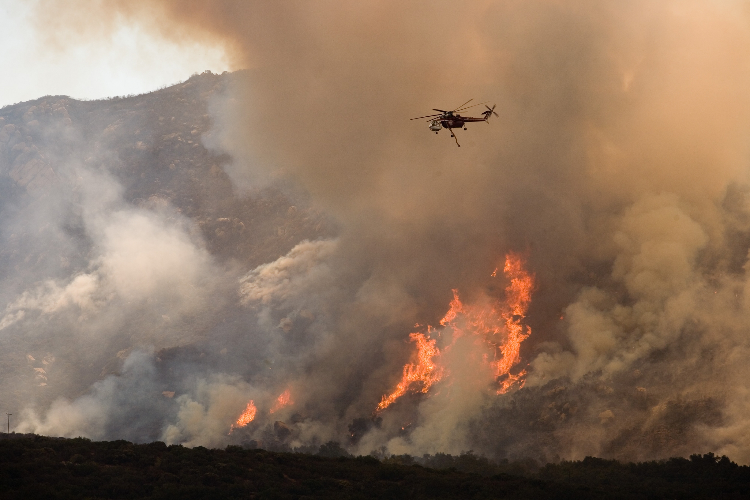 A helicopter flies closely over a wildfire on a mountain. You can see orange flames amongst a large cloud of white smoke. 