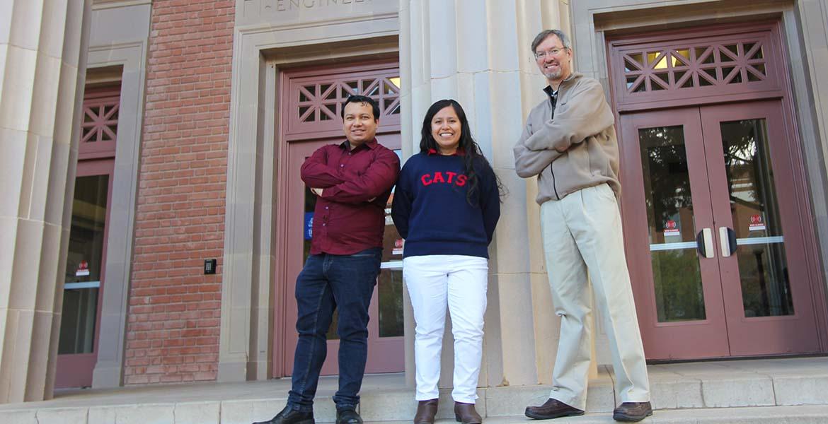 Two students stand with a professor for a photo in front of an academic building. 