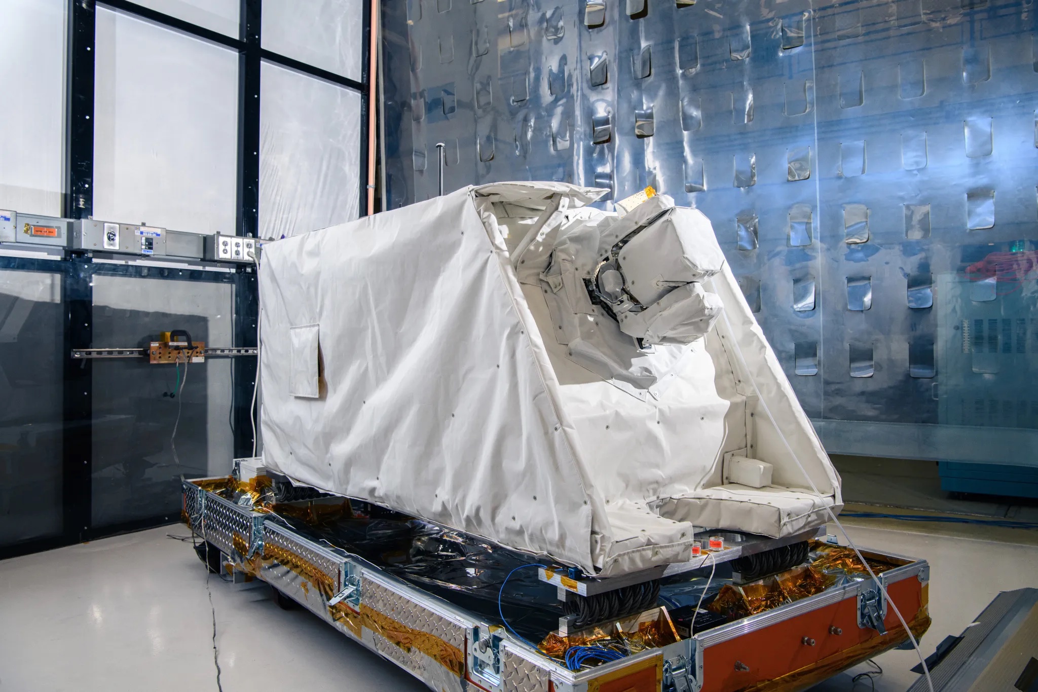 A refrigerator-sized structure with a white covering sits on a platform in a cleanroom. 