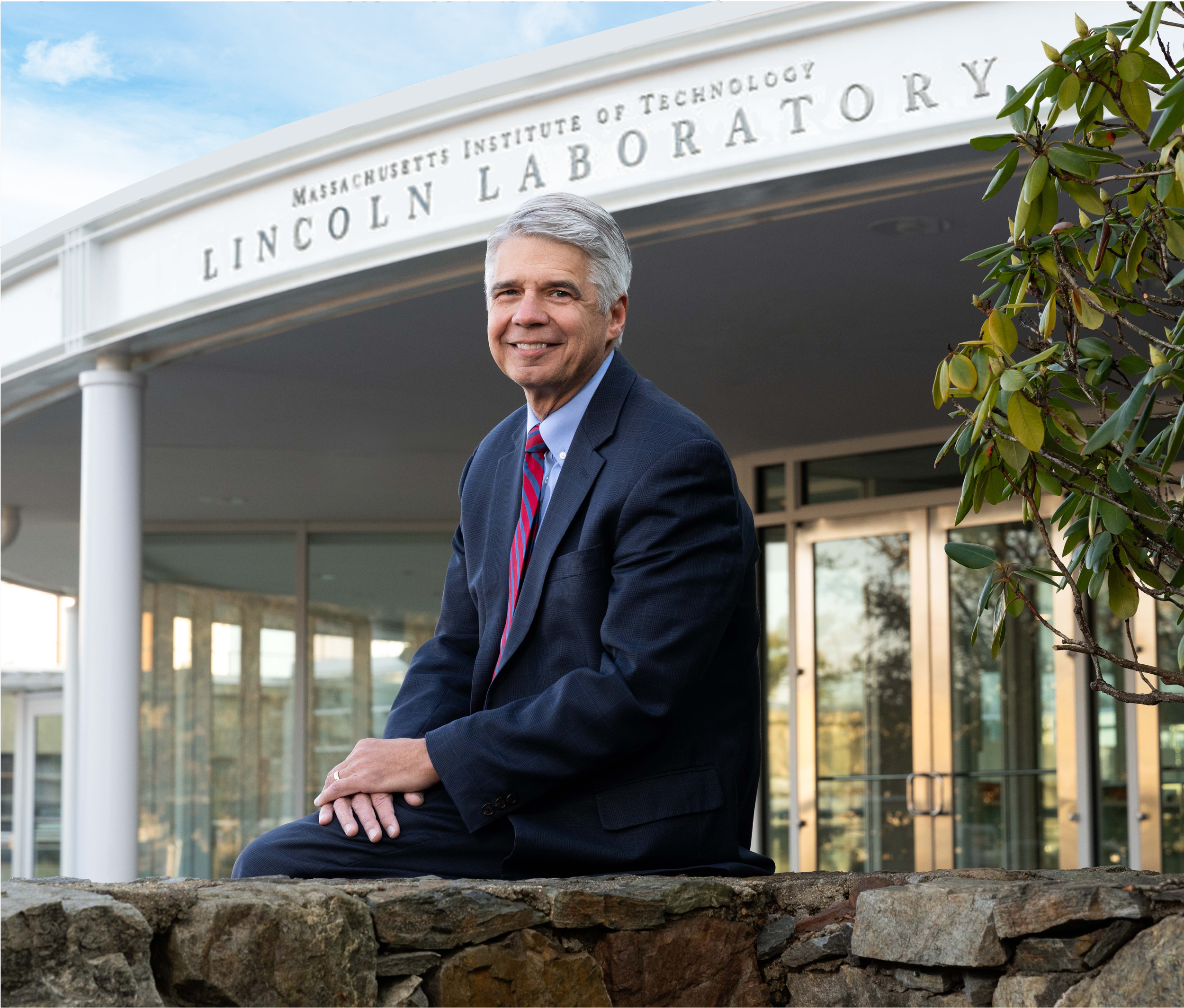 Eric Evans sits outside on a stone wall, in front of the Lincoln Laboratory's main entrance