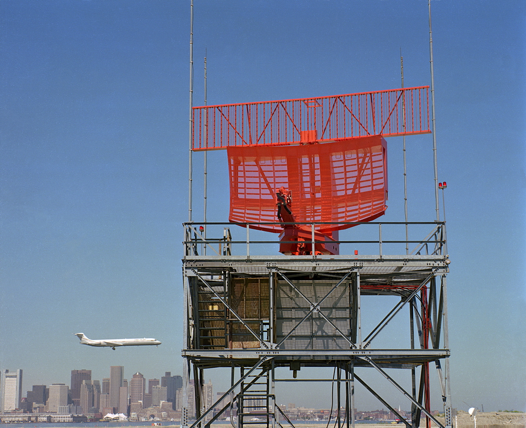 An exterior photo showing a large orange radar antenna. In the background, you see a low-flying commercial aircraft with the skyline of Boston behind that.