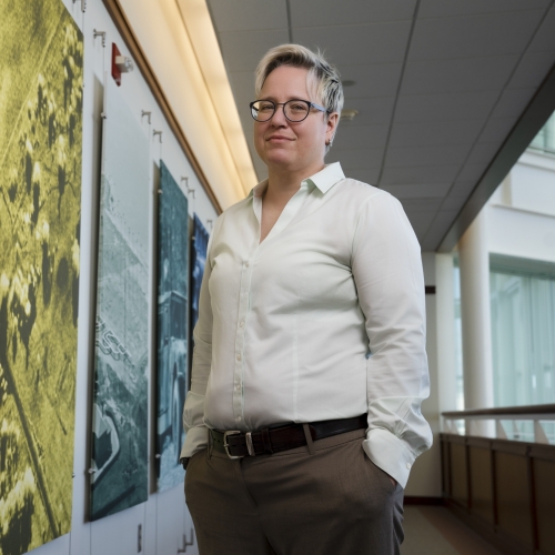 Molly Crane poses for a photo, smiling with her hands in her pockets, in a hallway. To her right is a wall with photos on it, one orange, then yellow, then blue. To her left is an indoor balcony.