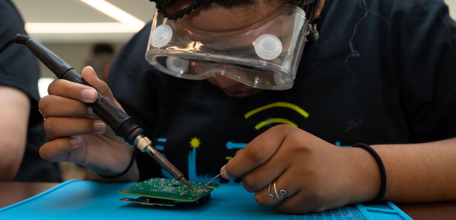 Hogan is sitting at a table wearing a blue LLRISE t-shirt and goggles as she solders a circuit.