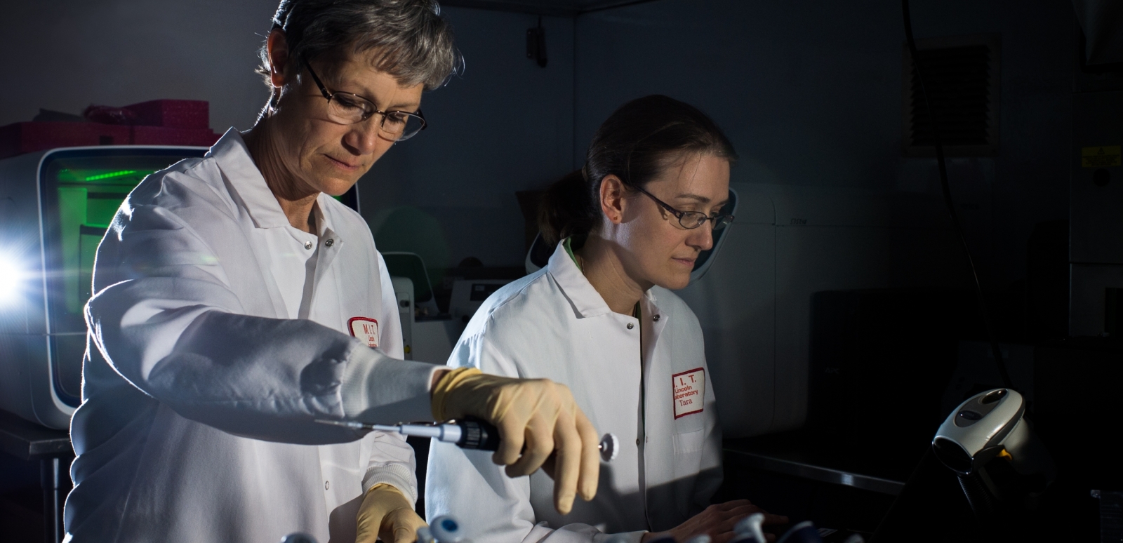 Two women in white lab coats standing with vials in foreground.