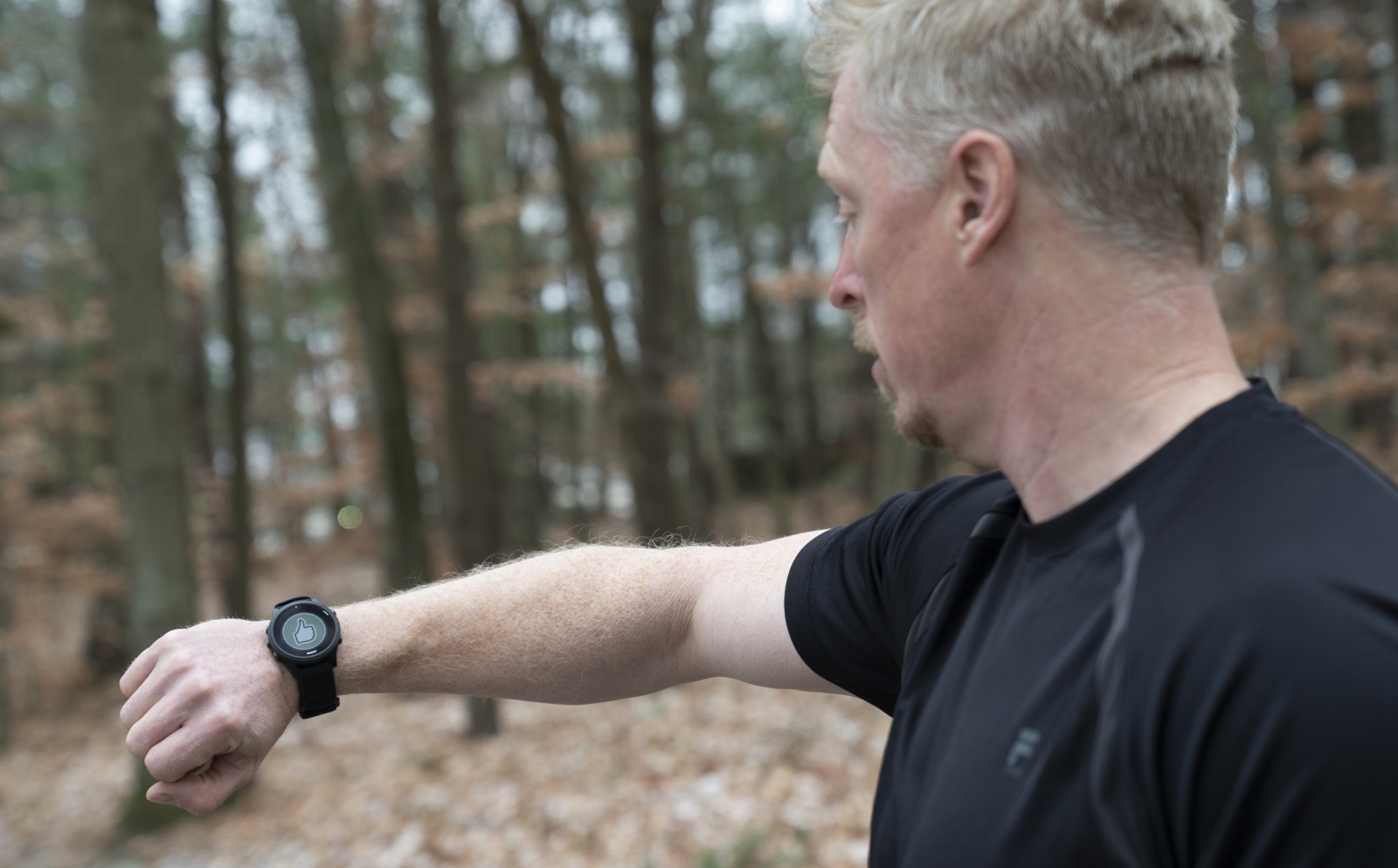 A man in black t-shirt looks at a smartwatch on his wrist, which is displaying a green circle with a thumbs up sign. The man is outside, with trees and leaves in the background.