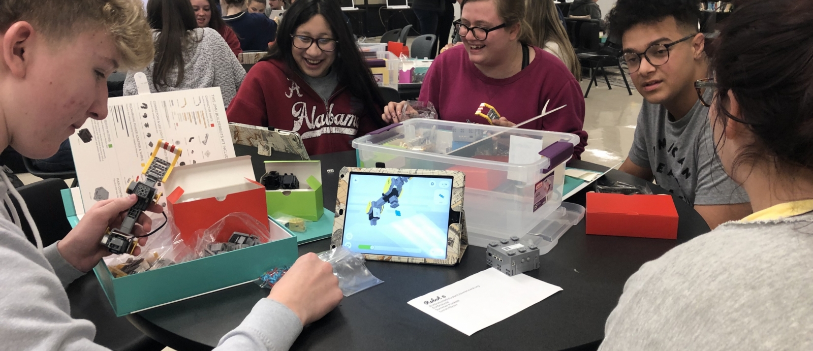 A group of five high school students sit at a table, smiling, working together to assemble a robot out of what look to be lego-like parts. 