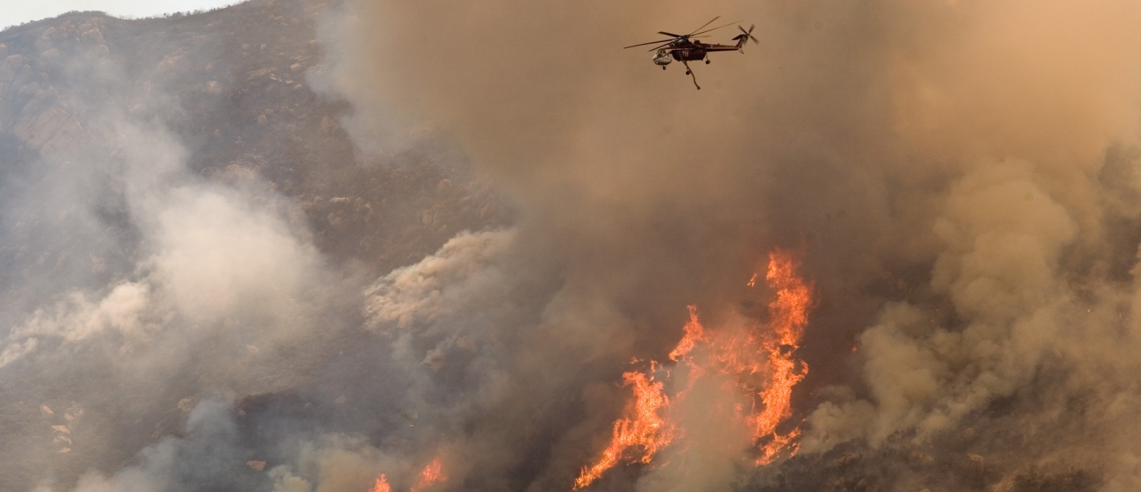 A helicopter flies closely over a wildfire on a mountain. You can see orange flames amongst a large cloud of white smoke. 