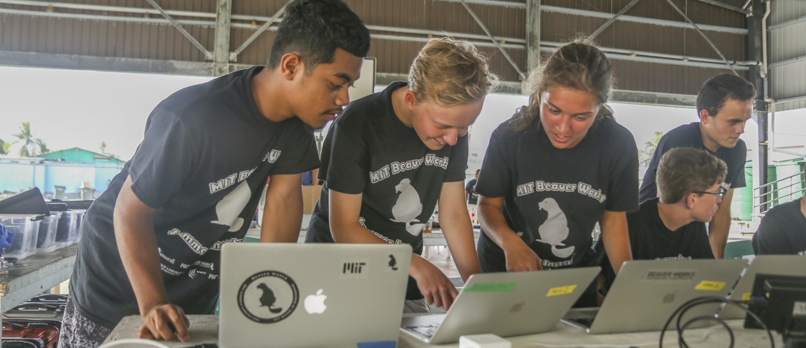 Three students stand together at a table, looking down each at a laptop, discussing. Two other students are in the background. The building they are in looks like an aircraft hanger. 