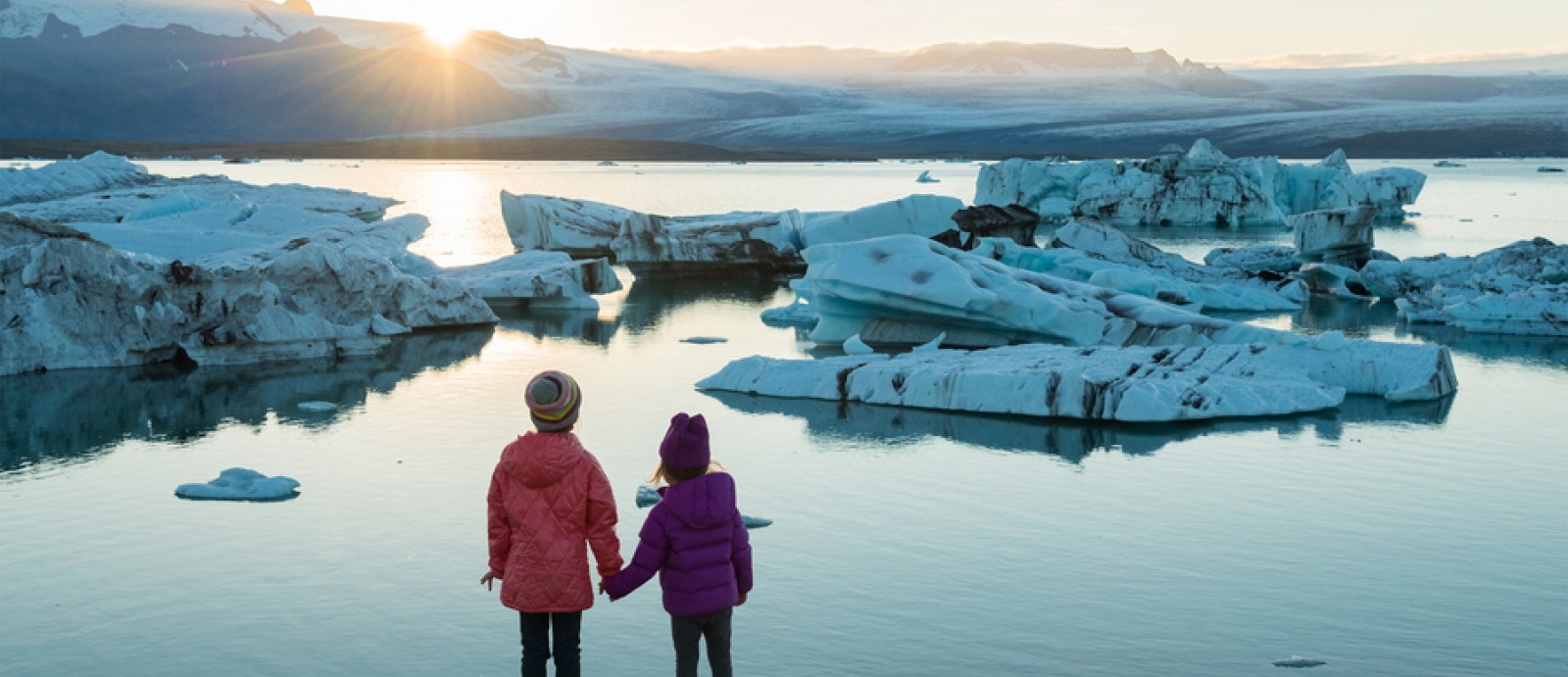 Two young girls hold hands while looking over glaciers and water. 