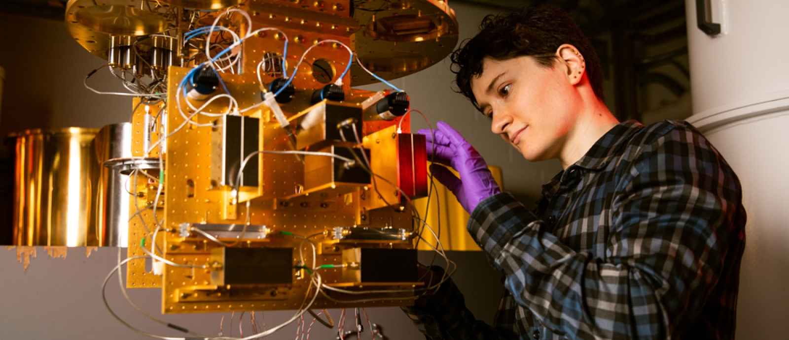 Alex Green stands at a dilution refrigerator, a gold/metal contraption. 