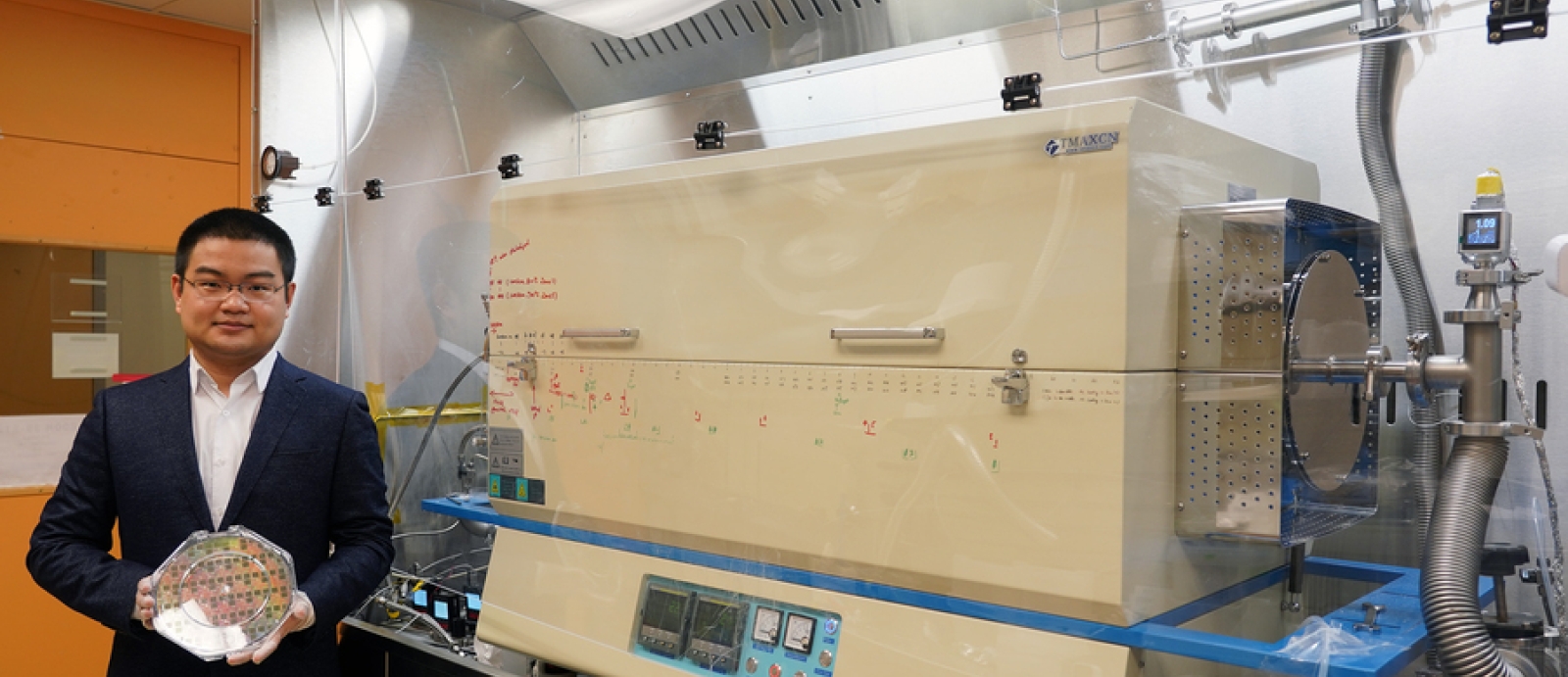 A researcher holds a wafer, while standing in front of a material deposition machine.