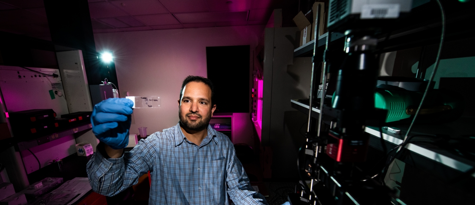 Siddharth Samsi holds a microscope slide containing a brain tissue sample. The sample will be imaged using the hyperspectral and quantitative phase imager system, at right, set up in the Biophotonic, Electric, Acoustic, and Magnetic Measurement Lab.