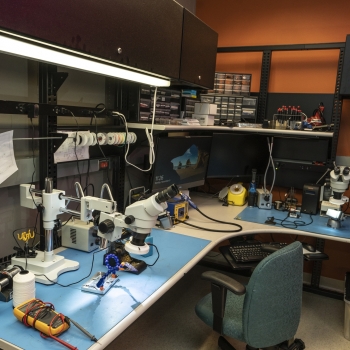 Photo of a desk that has a variety of equipment on it, including microscopes and soldering equipment.