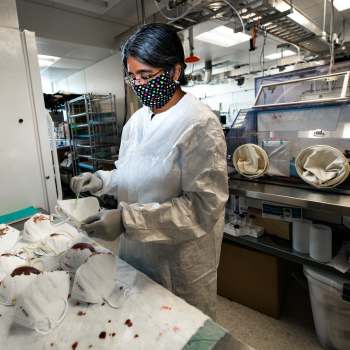 A woman in a lab coat and mask tests other masks for sythentic blood seepage -- a table of masks are in front of her with fake blood stains on them. 