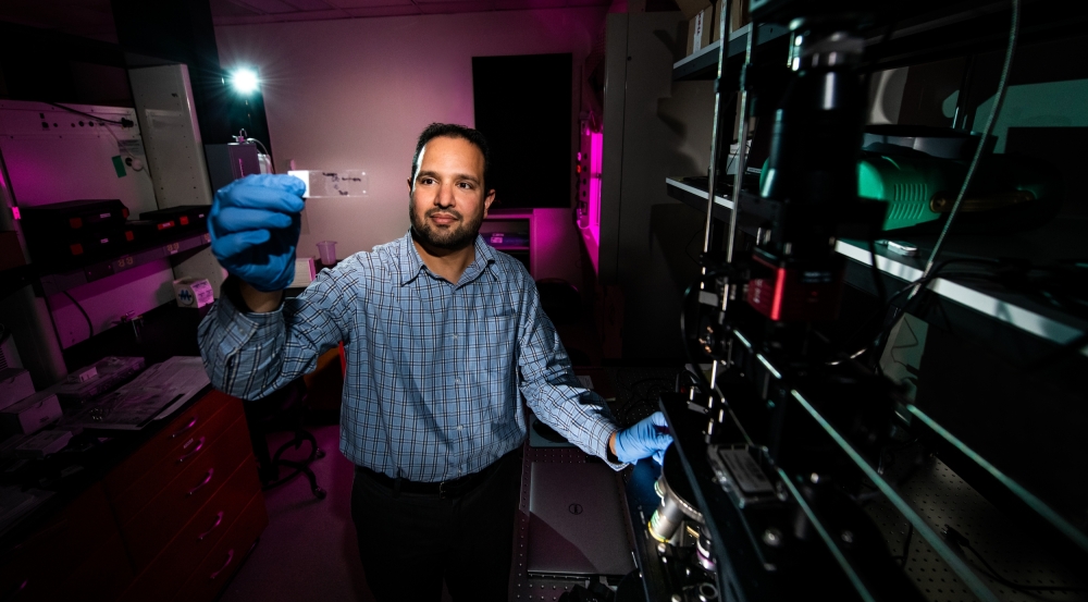 A researcher stands in a lab next to a microscope holding up a glass slide containing human tissue samples.