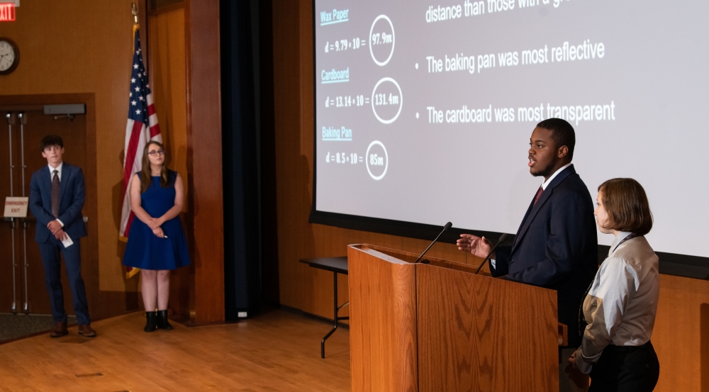 Four high school students stand on an auditorium stage giving a presentation. 