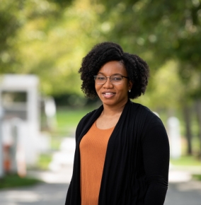 Shamaria Engram stands and smiles for a photo outside; she's wearing an orange top with a black sweater, green trees are out of focus in the background. 