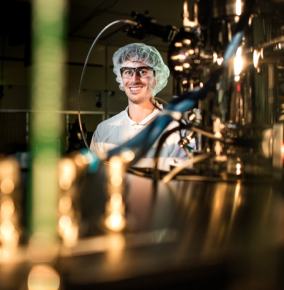 A photo of Dan in a quantum laboratory. He is wearing a hairnet, glasses, and a lab coat. Metal tubes and wires are in the background and foreground.