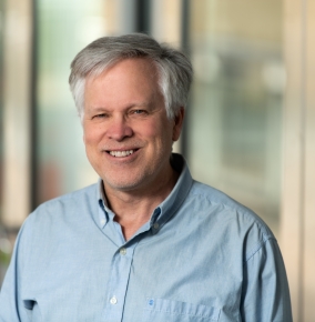 Jim Williamson poses for a photo, smiling, with a wall of windows in the background.