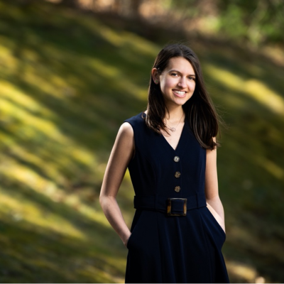 A photo of a woman standing outside, standing with her hands in her pockets and smiling. The background is a green field. 