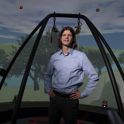 A photo of Brian Baum standing on a treadmill with support beam surrounding him. In the background is a screen showing a cloudy sky and trees. He is standing is a virtual reality dome.