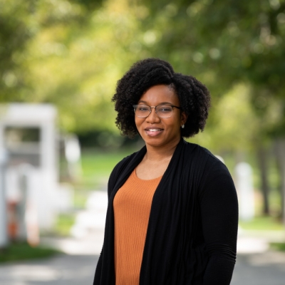 Shamaria Engram stands and smiles for a photo outside; she's wearing an orange top with a black sweater, green trees are out of focus in the background. 