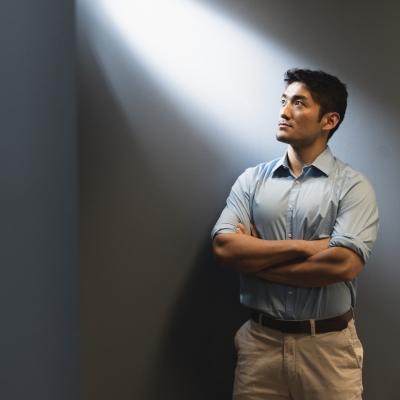 Yutai Zhou stands with his arms crossed, against a blue-grey wall. He looks up at light coming through a window.