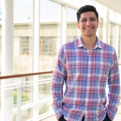 Gabriel Torres wears a plaid button-up, smiling for a photo in a sunlight hallway.