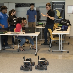 Students sitting around tables discuss the upcoming race surrounded by laptops. Two RC cars are stationed on the floor.