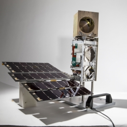 A small satelitte photographed against a white background. About , about 10 cm X 10 cm X 36 cm in size, it consists of a small solar panel array, connected to the satellite bus standing upright, topped with a golden cube. 