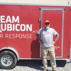 A photo of man in a khakis, a grey tshirt, and red hat standing for a photo in front of a red trailer, with the words Team Rubicon on it. 