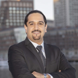 A photo of Hamed Okhravi. He is wearing a suit, with his arms crossed, with city buildings in the background. 