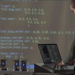 A researcher stands on a laptop. In front of him are a few small radios, and a large projector screen showing data. 