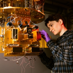 Alex Green stands at a dilution refrigerator, a gold/metal contraption. 