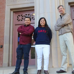 Two students stand with a professor for a photo in front of an academic building. 