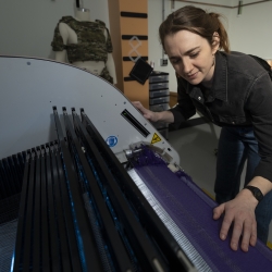 Erin Doran, a textile specialist, is photographed next to a textile weaving machine, with her hands on a fabric being woven. 