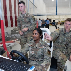 Three cadets are photographed; one is operating a steering wheel, controlling a robot off screen. The other two look on.