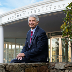 Eric Evans sits outside on a stone wall, in front of the Lincoln Laboratory's main entrance