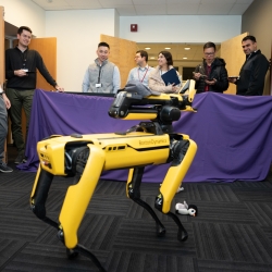 A yellow robotic dog stands in the foreground, with staff looking at it in the background.
