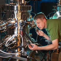 a researcher looks through a small window on a large metal multichambered molecular beam epitaxy tool.