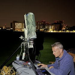 A person sits in front of a laptop. A telescope is next to him, pointed up. They are outside, with the city skyline behind them.
