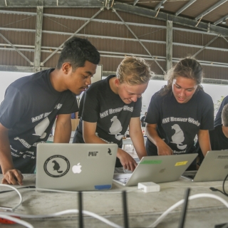 Three students stand together at a table, looking down each at a laptop, discussing. Two other students are in the background. The building they are in looks like an aircraft hanger. 