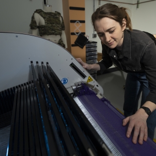 Erin Doran, a textile specialist, is photographed next to a textile weaving machine, with her hands on a fabric being woven. 