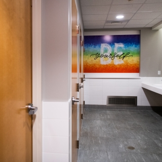 A photo showing the interior of the all-gender bathroom. On the left-hand side are doors to the hard-walled stalls, in the middle is a sign saying "Be Yourself," and to the right are sinks.
