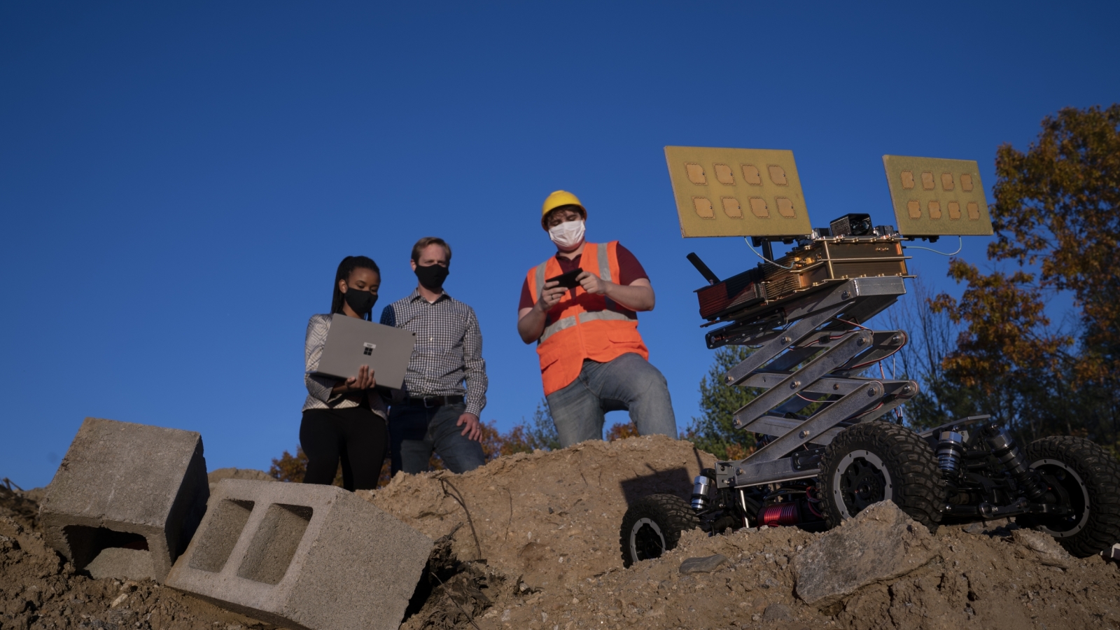 three people stand on a big dirt pile, outside, with blue sky in the background. One researcher is holding a laptop, which another looks at. The third person is holding a remote, controlling a robotic vehicle also on the dirt pile. 