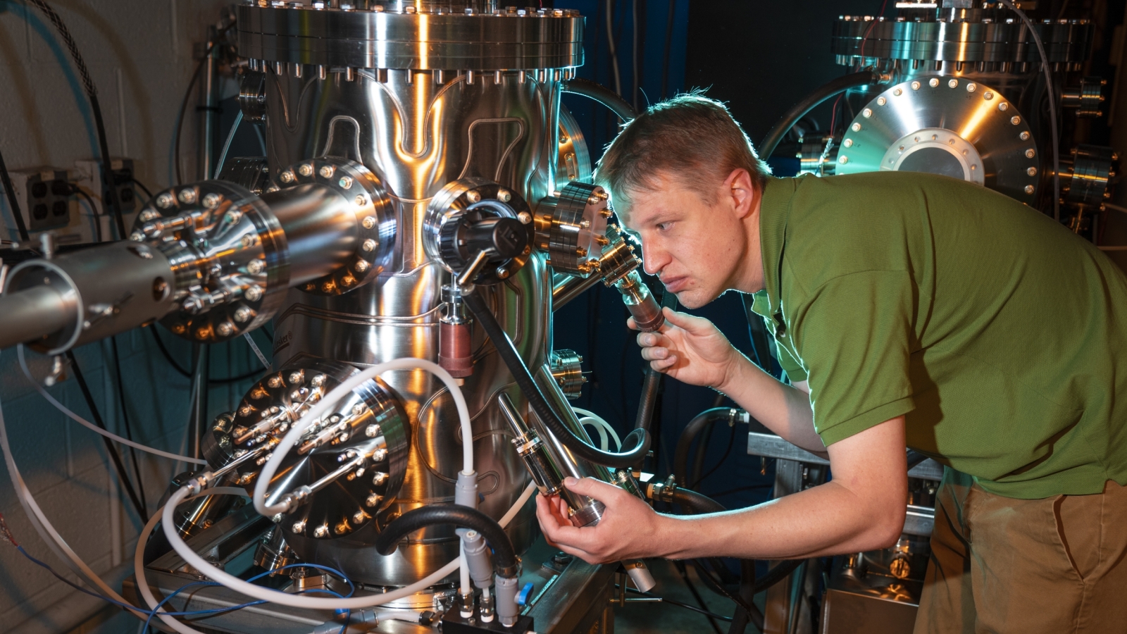 a researcher looks through a small window on a large metal multichambered molecular beam epitaxy tool.